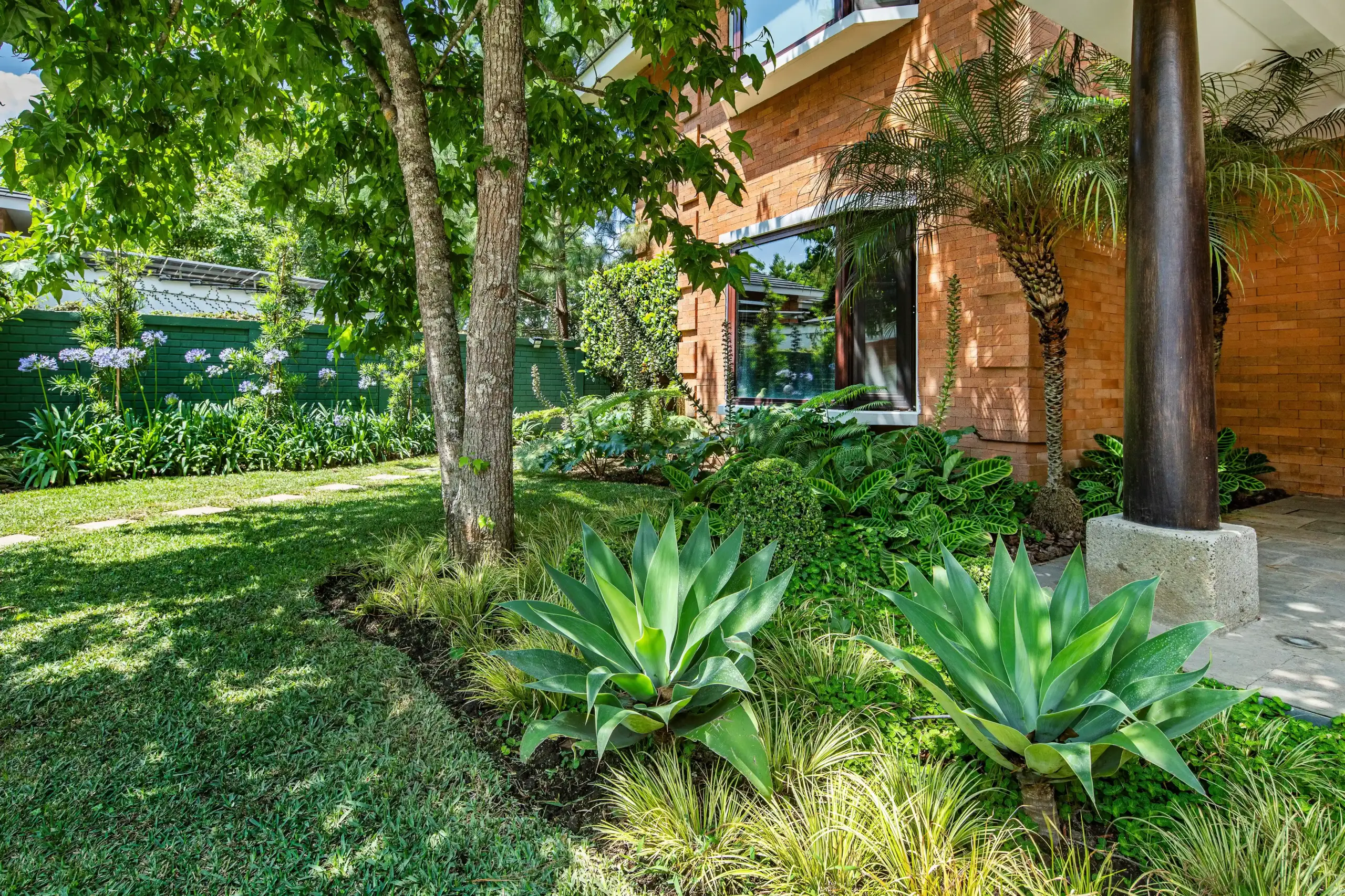 Jardín residencial con árboles, agaves grandes, césped y columnas de madera en casa de ladrillo en Guatemala